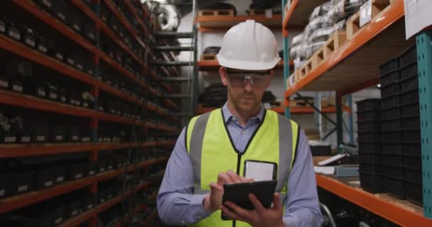 Un ouvrier d'usine de race blanche dans une usine fabriquant des équipements hydrauliques, portant un casque, des lunettes de sécurité et un gilet pare-balles, utilisant un ordinateur tablette, marchant entre les étagères dans un entrepôt de stockage, au ralenti .