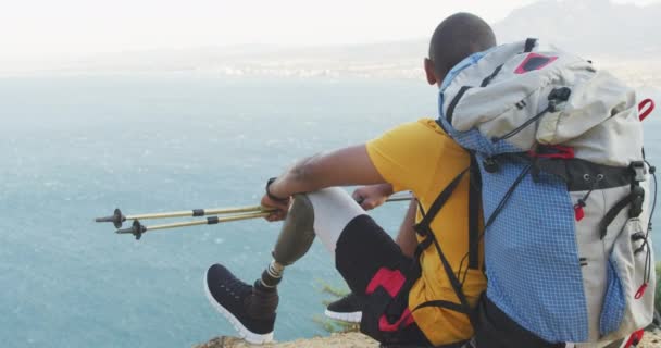 健康的 残疾的混合型赛车手 喜欢登山 坐在岩石上 拿着北欧手杖 在多云的天气里 慢吞吞地看着大海 — 图库视频影像