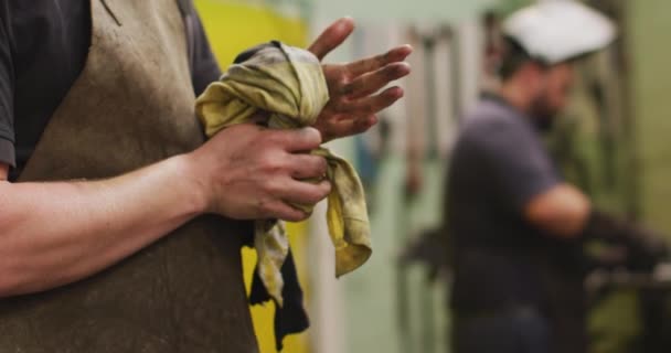 在一家生产液压设备的工厂里 一名白人男性工厂工人的中间部分 他用一块布洗手 另一个人在后面慢吞吞地干活 — 图库视频影像