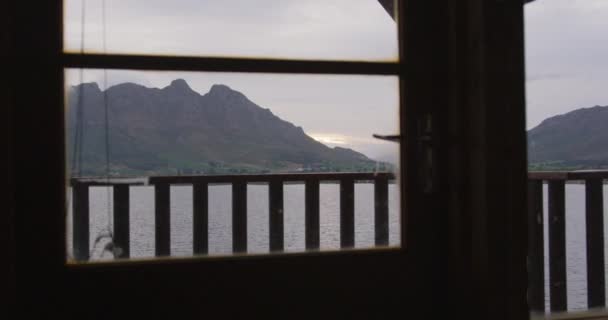 ログキャビンの窓から見える曇りの日の日没時の湖と山の眺めが遅い動き — ストック動画