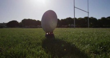 Kırmızı ve beyaz takım elbiseli, beyaz bir ragbi oyuncusu, yavaş çekimde bir oyun sahasında rugby topunu tekmeliyor.