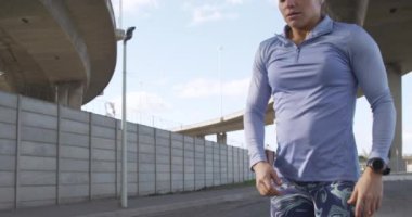 Güneşli bir günde şehir merkezinde egzersiz yapan, elleri dizlerinin üzerinde, yavaşça koştuktan sonra dinlenen, uzun siyah saçlı beyaz bir kadının yan görüntüsü..