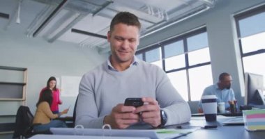 Kafkasyalı bir erkek iş adamının gündelik modern bir ofiste çalışırken, akıllı telefonunu ve laptopunu kullanarak arka planda yavaş çekimde iş arkadaşlarıyla birlikte masasında oturması düşük açılı bir bakış açısı.