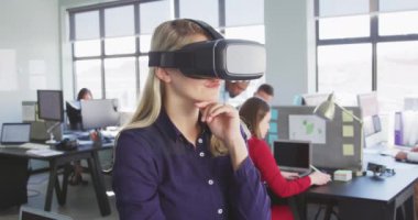Sarışın, beyaz, modern bir ofiste çalışan, VR kulaklık takan ve sanal interaktif ekrana dokunan, arka planda yavaş çekimde çalışan iş arkadaşlarının yan görünüşü.