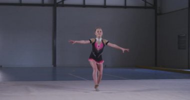 Odaklanmış genç beyaz kadın jimnastikçinin spor salonunda performans sergilemesi, kolları açık atlaması, ağır çekimde pembe ve siyah mayo giymesi..