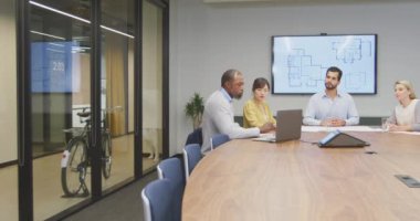 Gündelik modern bir ofiste çalışan Afrikalı Amerikalı bir erkek iş adamının yan görünüşü, bir dizüstü bilgisayar kullanarak ve bir toplantıda çeşitli meslektaşlarıyla konuşurken.