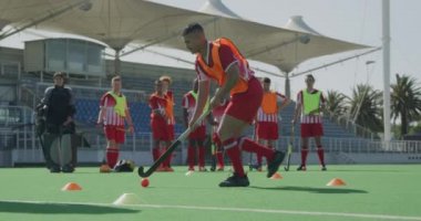 Melez bir erkek çim hokeyi oyuncusunun yan görüntüsü, maçtan önce antrenman, slalom topuyla hokey sopasıyla koşması, stadyum ve takım arkadaşlarıyla güneşli bir günde arka planda, ağır çekimde koşması.