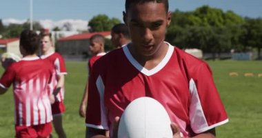 Kırmızı ve beyaz takım elbiseli genç bir rugby oyuncusunun portresi, bir oyun sahasında duruyor, bir ragbi topu tutuyor ve kameraya bakıyor, diğer oyuncular arka planda yavaş çekimde ısınıyor.