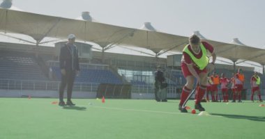 Kafkasyalı bir erkek çim hokeyi oyuncusunun yan görüntüsü, maçtan önce antrenman yapması, hokey sopasıyla topa vurması, güneşli bir günde arka planda takım arkadaşlarıyla birlikte yavaş çekim yapması.