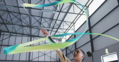 Salonda performans sergileyen, kurdeleyle egzersiz yapan, kurdeleyi kafasının üstünde döndüren, yavaş çekimde çok renkli mayo giyen, odaklanmış genç bayan jimnastikçinin düşük açılı yan görüntüsü..