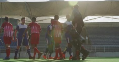 İki takımın çim hokeyi oyuncularından oluşan çok ırklı bir grubun yan görüntüsü. Güneşli bir günde hokey sopalarıyla ağır çekimde el sıkışıyorlar.