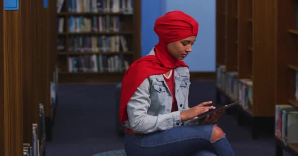 一名亚洲女学生穿着红色头巾在图书馆学习 坐着用平板电脑 穿着休闲装 — 图库视频影像