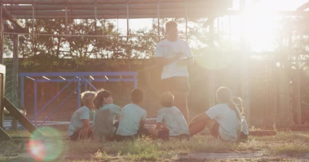 在阳光灿烂的日子里 看到一群快乐的白人男孩女孩和一个白人男孩一起在新兵训练营 孩子们坐在那里 教练慢慢地指导他们 他们都穿着绿色的T恤衫 — 图库视频影像