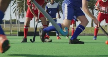 Kafkasyalı genç bir erkek çim hokeyi oyuncusunun yan görüntüsü. Hokey maçı sırasında sahada topla koşuyor. Rakip takım arka planda güneşli bir günde, ağır çekimde.