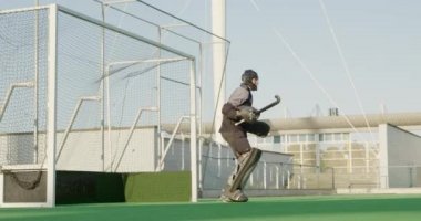 Kafkasyalı bir erkek çim hokeyi kalecisinin yan görüntüsü, hokey sahasında hokey sopasıyla topu durdurmak için dalması, takım arkadaşları ve rakipleriyle güneşli bir günde arka planda, yavaş çekimde