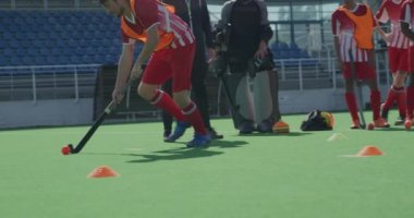 Kafkasyalı bir erkek çim hokeyi oyuncusunun yan görüntüsü, maçtan önce antrenman yapması, hokey sopasıyla topa vurması, güneşli bir günde arka planda takım arkadaşlarıyla birlikte yavaş çekim yapması.