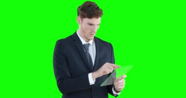 Beyaz gömlek ve takım elbise giyen yakışıklı beyaz adam yeşil arka planda şeffaf ekran tabletini kullanıyor..