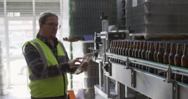 Yüksek görüş mesafesi olan beyaz bir adam, bira fabrikasında çalışıyor, bira şişelerini kontrol ediyor, bilgisayar tabletini kullanıyor, Afrikalı Amerikalı bir adam çalışıyor, şişeleri yavaş çekimde kontrol ediyor..