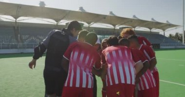 Çok ırklı bir grup genç çim hokeyi oyuncusu ve beyaz erkek çim hokeyi koçu, maçtan önce motivasyon sağlıyorlar, el ele tutuşuyorlar, güneşli bir günde sahada yavaş çekimde etkileşime geçiyorlar.