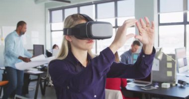 Sarışın, beyaz, modern bir ofiste çalışan, VR kulaklık takan ve sanal interaktif ekrana dokunan, arka planda yavaş çekimde çalışan iş arkadaşlarının yan görünüşü.