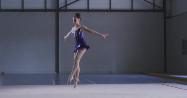 Spor salonunda performans sergileyen beyaz kadın jimnastikçinin yan görünüşü, kolları açık atlamak, ağır çekimde mor ve beyaz mayo giymek..