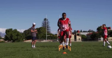Kırmızı ve beyaz takım elbiseli çok ırklı genç bir erkek rugby takımının ön görüntüsü. Oyun sahasında antrenman yapıyorlar, atlıyorlar ve yanlarında ağır çekimde koçları var.