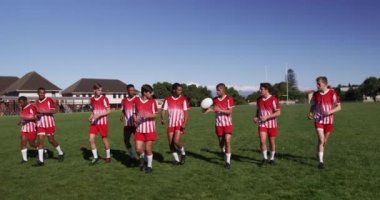 Kırmızı ve beyaz takım elbiseli, futbol sahasında antrenman yapan ve ağır çekimde ragbi topu fırlatan çok ırklı genç bir rugby takımı ön planda.