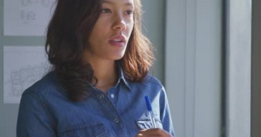 Modern bir ofiste çalışan, pencerenin kenarında duran, kalemle çenesine dokunan ve güneşli bir günde yavaşça gülümseyen melez bir iş kadınının ön görüntüsü.