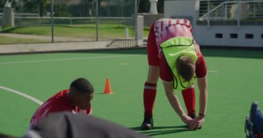Beyazlı bir genç hokey oyuncusunun maçtan önce antrenman yapmasını, çim hokeyi sahasında antrenman yapmasını bacaklarını esnetmesini, eğilmesini, melez takım arkadaşının güneşli bir günde önünde oturmasını.