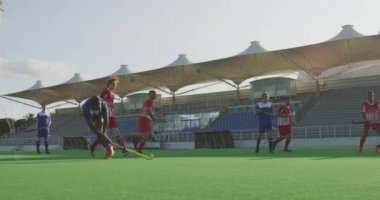 Çok ırklı iki genç çim hokeyi takımının yan görüntüsü, çim hokeyi oyunu oynuyorlar, kaleci şutu savunuyor ve takım arkadaşları ona tezahürat yapıyor, güneşli bir günde, ağır çekimde.