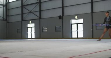 Genç melez kadın jimnastikçinin spor salonunda performans sergilemesi, bir çemberle egzersiz yapması, zıplaması, ağır çekimde siyah mayo giymesi..