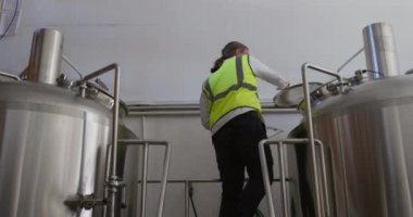 Bir bira fabrikasında çalışan beyaz bir adam, yüksek görünürlüklü bir yelek giyiyor, birayı inceliyor ve tankın içine bakıyor, yavaş çekimde notlar alıyor..