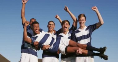 Mavi ve beyaz takım elbiseli bir grup genç rugby oyuncusunun önünde zaferi kutluyor, takım arkadaşlarından birini taşıyor ve kollarıyla havada tezahürat yapıyor, yavaş çekimde bir oyun sahasında duruyor.