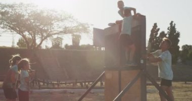 Bir grup beyaz erkek ve kızın, güneşli bir günde acemi birliğindeki beyaz bir erkek koçun, yeşil tişörtler giyerek duvarın üzerinden tırmanmasının yan görüntüsü.