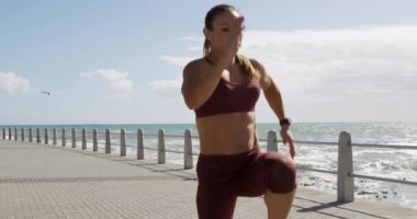 Mavi gökyüzü ile güneşli bir günde, deniz kenarında uzun siyah saçlı sportif bir beyaz kadın, yavaş çekimde koşuyor, zıplıyor ve tek bacağını kaldırıyor..