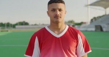 Kırmızı takım elbisesi giyen, hokey sahasında durup kameraya bakan, güneşli bir günde ağır çekimde gülümseyen melez bir hokey oyuncusunun portresi.