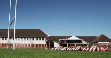 Kırmızı ve beyaz takım elbiseli, futbol sahasında antrenman yapan, arka arkaya mekik çeken ve yanında ağır çekimde koçu olan çok ırklı genç rugby oyuncularından oluşan bir erkek takımının yan görüntüsü.
