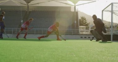 Çok ırklı iki genç çim hokeyi takımının yan görüntüsü, çim hokeyi oyunu oynuyorlar, oyunculardan biri hokey sopasıyla topa vuruyor ve kaleci, güneşli bir günde, ağır çekimde şutu bacağıyla savunuyor.