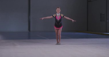 Odaklanmış genç beyaz kadın jimnastikçinin spor salonunda performans sergilemesi, kolları açık atlaması, ağır çekimde pembe ve siyah mayo giymesi..