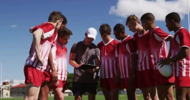 Kafkasyalı bir erkek rugby koçunun, çok ırklı rugby oyuncularından oluşan genç bir erkek takımla, takım elbiselerini giymiş ve oyun sahasında ağır çekimde ayakta durduğunu görüyoruz.