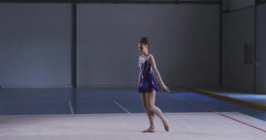 Spor salonunda performans sergileyen beyaz kadın jimnastikçinin yan görünüşü, kolları açık atlamak, ağır çekimde mor ve beyaz mayo giymek..