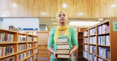 Sarı tesettürlü Asyalı bir kız öğrenci kütüphanede ders çalışıyor, kitaplıkların arasında yürüyor ve bir yığın kitap tutuyor, günlük giysiler giyiyor.