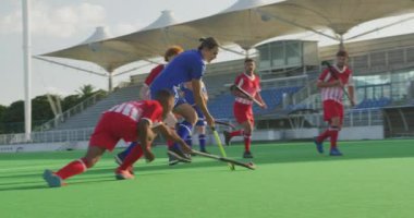 Çok ırklı iki genç çim hokeyi takımının yan görüntüsü, çim hokeyi oyunu oynuyorlar, oyunculardan biri defansta koşuyor ve topu takım arkadaşına paslıyor, o da güneşli bir günde, ağır çekimde topu atamıyor.