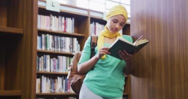 Sarı tesettürlü Asyalı bir kız öğrenci kütüphanede ders çalışıyor, kitaplıklara yaslanıyor, kitap okuyor, günlük giysiler giyiyor.