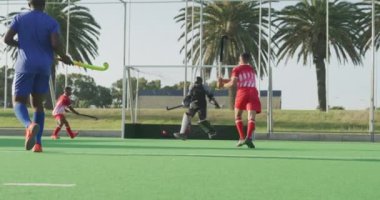 Çok ırklı iki genç çim hokeyi takımının yan görüntüsü, çim hokeyi maçı, oyunculardan biri hokey sopasıyla topa vuruyor ve kaleci, güneşli bir günde, ağır çekimde, bacağıyla şutu savunamıyor.