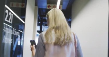 Kafkasyalı bir kadın iş kadınının modern bir ofisin koridorunda akıllı telefon kullanarak yürüdüğüne dair arka bakış açısı.