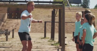 Güneşli bir günde Acemi Birliğindeki beyaz bir erkek koç ve beyaz bir grup mutlu erkek çocuğun yan görüntüsü. Bir koç çocuklara yeşil tişörtler giymelerini söylüyor.