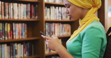Sarı tesettürlü gülümseyen Asyalı bir kız öğrenci kütüphanede çalışıyor, ayakta duruyor ve akıllı telefon kullanıyor, günlük kıyafetler giyiyor.