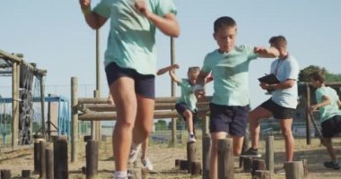 Güneşli bir günde Acemi Birliğindeki beyaz bir erkek koç, yeşil tişörtlü ve ağır çekimde ahşap kazıkların üzerinde koşuyor.