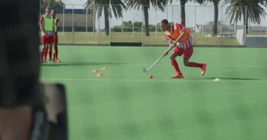 Kafkasyalı bir erkek çim hokeyi kalecisi, hokey sahasında hokey sopasıyla topu durdurmak için dalırken, arka planda arkadaşlarıyla güneşli bir günde, ağır çekimde...